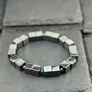 May include: A bracelet composed of metallic grey cube-shaped beads arranged in a circular pattern. The beads feature a smooth, reflective surface and are separated by small, round metallic spacers, creating a uniform and structured appearance.