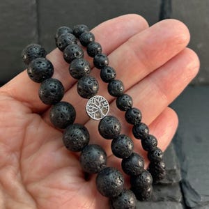 May include: A beaded bracelet featuring porous black lava stone spheres and a circular silver-toned charm depicting a tree. The bracelet is constructed with a series of round, textured volcanic rock beads that vary slightly in size, centred by the metallic tree emblem.