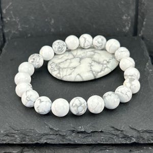 May include: A white and grey beaded bracelet with a matching oval stone. The bracelet features round, white beads with grey marbling. The oval stone has a similar white and grey pattern. The bracelet is displayed on a dark grey surface.