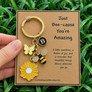 Peut inclure: Un porte-clés en métal doré avec une marguerite jaune, un papillon jaune, une abeille à strass et un médaillon noir avec la lettre B. Le porte-clés est fixé sur une carte beige portant le texte Just Bee-cause You're Amazing.