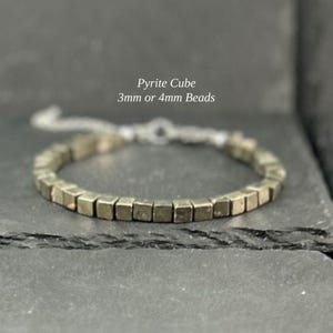 May include: A bracelet composed of metallic gold-toned pyrite cube beads. The text Pyrite Cube 3mm or 4mm Beads is visible above the bracelet. The jewellery features a silver-toned metal chain extension with a clasp mechanism for adjustable sizing.