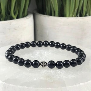 May include: A stretch bracelet composed of smooth, round, glossy black beads. A single circular silver-toned metal charm featuring an embossed cross design is positioned centrally among the black beads, creating a focal point on the accessory.