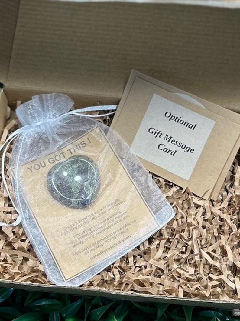 May include: A heart-shaped green stone inside a white organza drawstring bag, placed on a card with the text YOU GOT THIS! and a description of Dragon's Blood stone, alongside a separate card labeled Optional Gift Message Card.