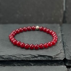 May include: A red beaded bracelet with a silver accent bead. The bracelet is made of small, round, polished red beads. The bracelet is displayed on a dark gray, textured surface.