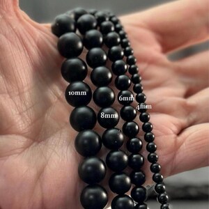 May include: Four strands of matte black spherical beads arranged by size. The strands are labeled with text indicating diameters of 10mm, 8mm, 6mm, and 4mm. The beads have a smooth, uniform finish and are aligned vertically to show the size progression.