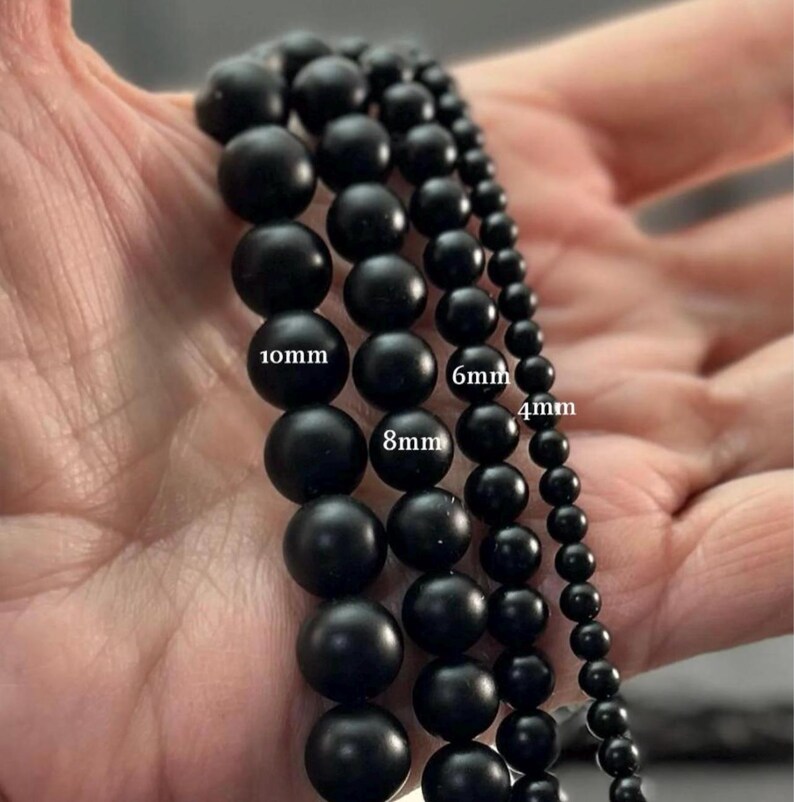May include: Four strands of matte black spherical beads arranged by size. The strands are labeled with text indicating diameters of 10mm, 8mm, 6mm, and 4mm. The beads have a smooth, uniform finish and are aligned vertically to show the size progression.