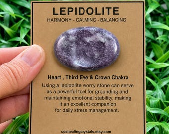 May include: An oval-shaped purple lepidolite worry stone mounted on a rectangular tan display card. The card features the text LEPIDOLITE, HARMONY - CALMING - BALANCING, Heart, Third Eye & Crown Chakra, and descriptive text about the stone.