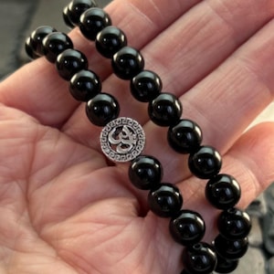 May include: Two stretch bracelets composed of smooth, round, polished black beads. Each bracelet features a single circular silver-toned metal charm with a Greek key border pattern surrounding a central Om symbol, creating a uniform look.