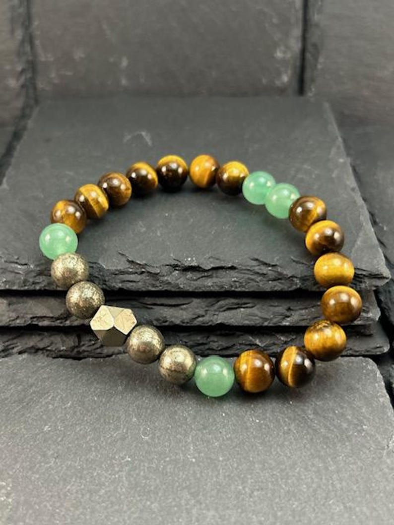 May include: A beaded bracelet featuring round tiger eye beads in shades of brown and gold, interspersed with smooth light green aventurine beads and metallic pyrite beads, including one faceted geometric pyrite bead in the center.
