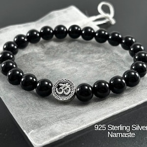 May include: A stretch bracelet featuring smooth, round black beads and a single circular silver-toned charm embossed with an Om symbol and a Greek key border. The text 925 Sterling Silver Namaste is printed in the lower right corner of the image.