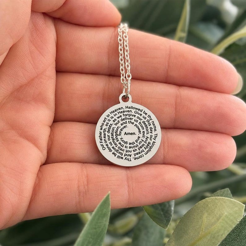 May include: A circular silver-coloured pendant necklace featuring the Lord's Prayer engraved in black text in a spiral pattern. The pendant is attached to a delicate silver-coloured chain link necklace, with the text ending in the word Amen at the centre.