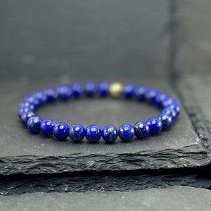 May include: A circular beaded bracelet composed of deep blue lapis lazuli stones with a single metallic gold-toned bead accent. The beads are uniform in size and arranged in a continuous loop, showcasing the natural texture of the blue stone.