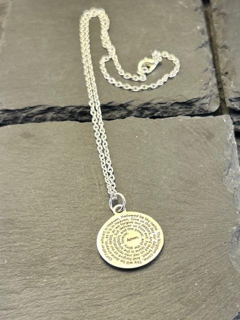 Peut inclure: Un collier argent&eacute; dot&eacute; d'un pendentif circulaire grav&eacute; du texte de la pri&egrave;re du Notre P&egrave;re en noir. Le pendentif est fix&eacute; &agrave; une fine cha&icirc;ne argent&eacute;e avec un fermoir mousqueton, formant un bijou simple et &eacute;l&eacute;gant pour le quotidien.