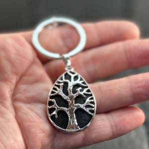 May include: A silver-toned keychain featuring a teardrop-shaped pendant. The pendant contains a black, textured, porous inlay overlaid with a polished silver-colored metal cutout depicting a tree with spreading branches and a trunk.