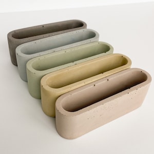 May include: Five concrete planters in various shades of green, gray, and brown. The planters are oval shaped and have a rough, textured surface.