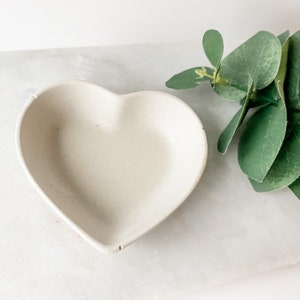 May include: A white heart-shaped ceramic dish on a white surface with green eucalyptus leaves.