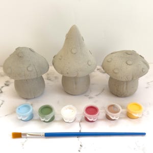 May include: Three gray concrete mushroom figurines with paint pots in blue, green, white, red, brown, and yellow, plus a blue-handled paintbrush. The mushrooms vary in size and shape, perfect for a DIY craft project.