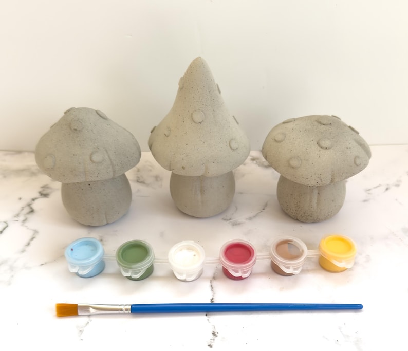 May include: Three gray concrete mushroom figurines with paint pots in blue, green, white, red, brown, and yellow, plus a blue-handled paintbrush. The mushrooms vary in size and shape, perfect for a DIY craft project.