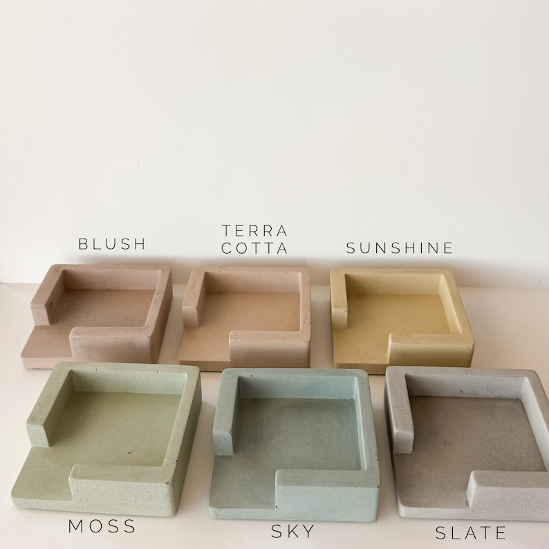 May include: A collection of six square concrete trays in various colors: blush, terra cotta, sunshine, moss, sky, and slate. Each tray has a raised edge on one side. The names of the colors are printed above each tray.