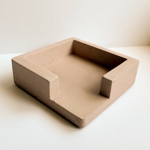 May include: A square, light brown concrete desk organizer with a rectangular opening. The organizer has a raised edge on three sides and a lower edge on the front, creating a slot. The item is designed to hold items like notepads or small office supplies.