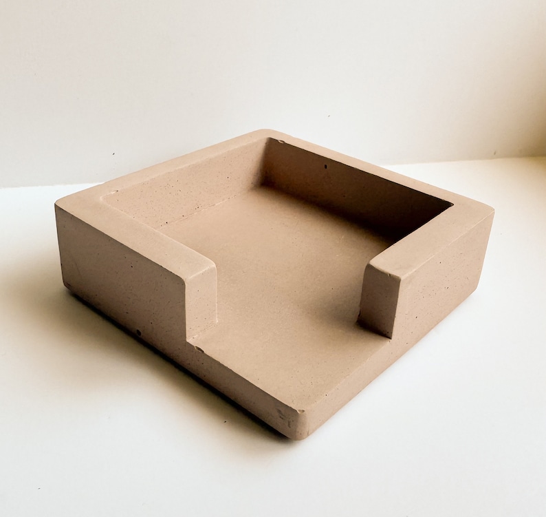 May include: A square, light brown concrete desk organizer with a rectangular opening. The organizer has a raised edge on three sides and a lower edge on the front, creating a slot. The item is designed to hold items like notepads or small office supplies.