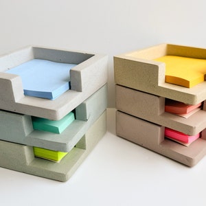 May include: Two sets of concrete desk organizers, each with three stacked trays. One set holds blue, green, and yellow sticky notes, while the other holds yellow, orange, and pink notes. The organizers have a modern, minimalist design.