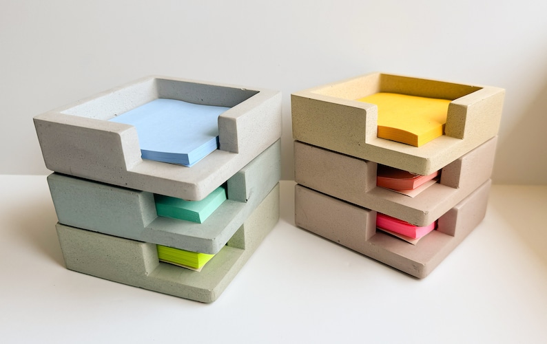 May include: Two sets of concrete desk organizers, each with three stacked trays. One set holds blue, green, and yellow sticky notes, while the other holds yellow, orange, and pink notes. The organizers have a modern, minimalist design.