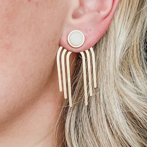Op de afbeelding: Goudkleurige oorbellen met een witte, cirkelvormige stud en een trapsgewijs ontwerp van verticale staven. De oorbellen worden getoond op een oor, met blond haar op de achtergrond. De oorbellen hebben een moderne, minimalistische stijl.