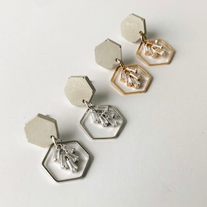 May include: Three pairs of earrings with hexagonal concrete tops and dangling geometric frames with crystal accents. The earrings are silver, gold, and silver.
