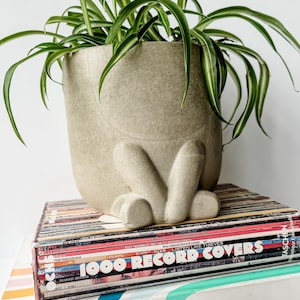 May include: A light gray ceramic planter shaped like a seated figure with a spider plant growing inside. The planter is sitting on a stack of vintage record albums with the text "1000 Record Covers" on the top album.