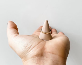 May include: A gold ring with a beaded design sits on a beige ceramic ring holder. The holder is shaped like a cone and is resting in the palm of a hand.