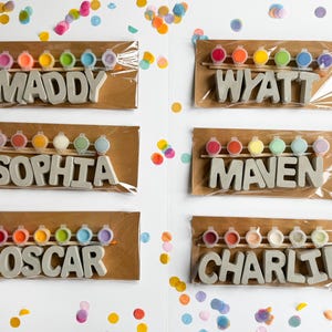 May include: Six personalized name kits with paint pots and wooden sticks. Each kit includes a name in gray letters, with names like Maddy, Wyatt, Sophia, Maven, Oscar, and Charlie. Colorful confetti surrounds the kits.