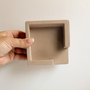 May include: A square, light brown concrete desk organizer with a rectangular cutout on one side. The organizer is held by a hand against a white background. The organizer is a neutral color.