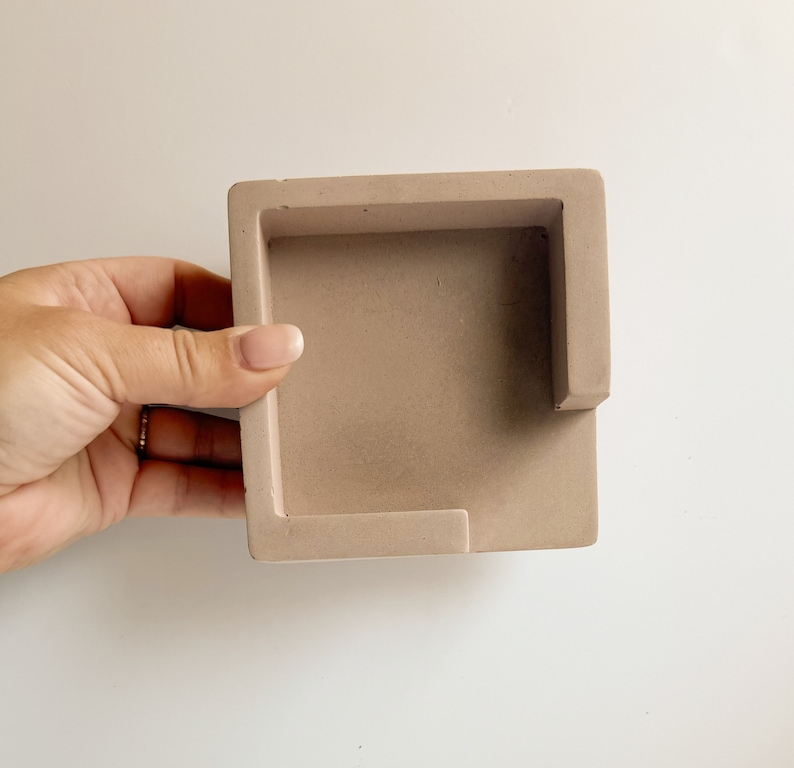 May include: A square, light brown concrete desk organizer with a rectangular cutout on one side. The organizer is held by a hand against a white background. The organizer is a neutral color.