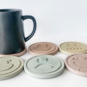 May include: Six round concrete coasters with different smiley faces. The coasters are in various shades of green, brown, and beige. The smiley faces include a winking smiley face, a smiley face with hearts for eyes, a sad smiley face, a smiley face with stars for eyes, and two smiley faces with squiggly lines for mouths.