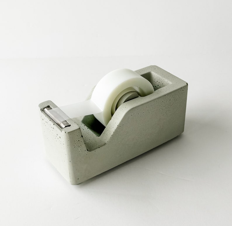 K&ouml;nnte beinhalten: Ein grauer Beton-Klebebandspender mit einer wei&szlig;en Klebebandrolle. Der Spender hat eine strukturierte Oberfl&auml;che und ein Metallmesser.