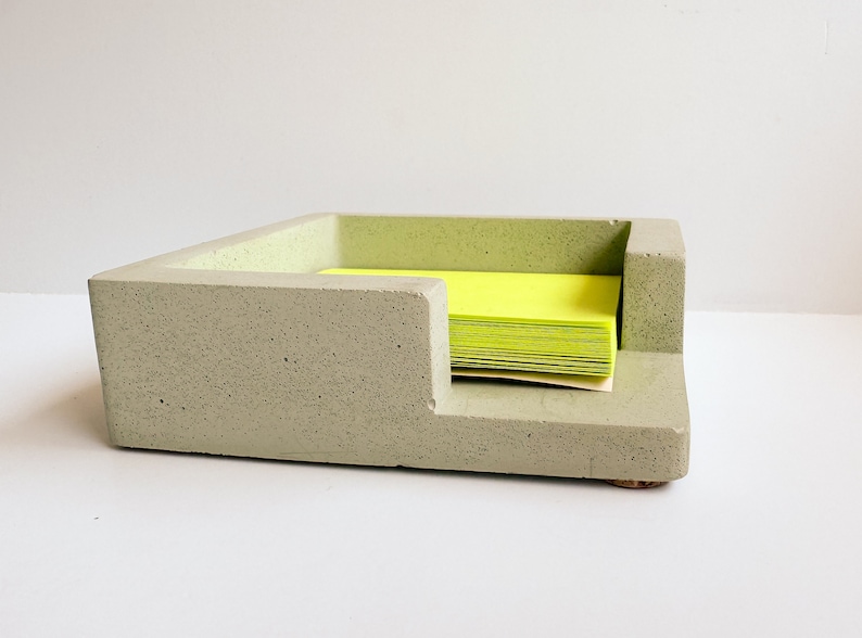 May include: A light green concrete desk organizer with a stepped design. The organizer holds a stack of bright yellow sticky notes. The desk accessory has a modern, minimalist aesthetic and is suitable for home or office use.