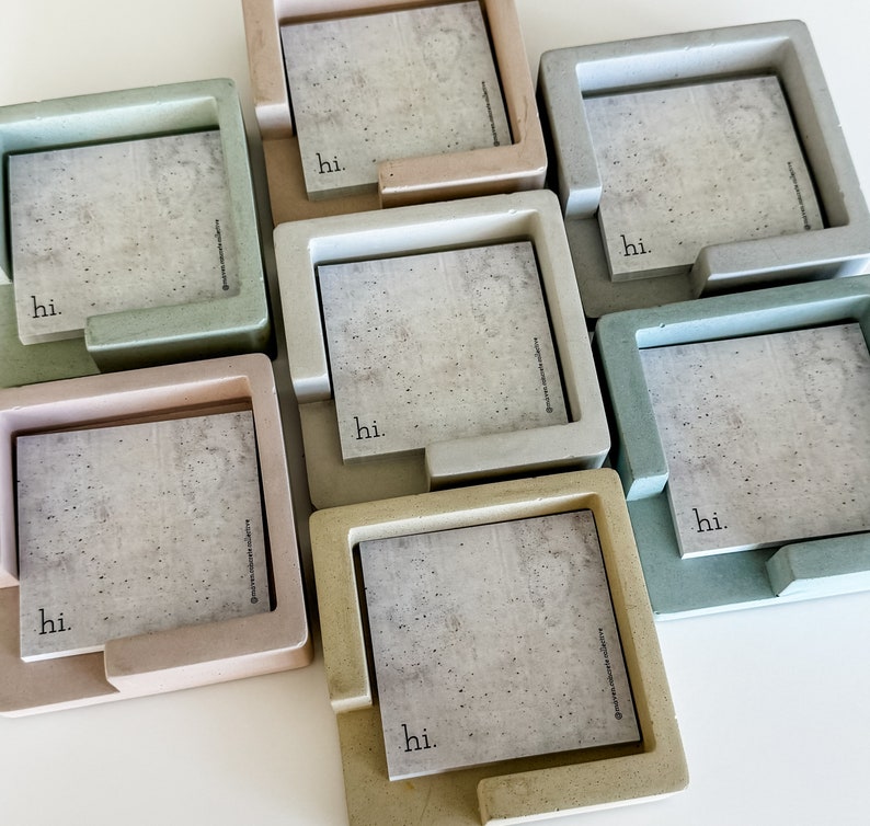 May include: A collection of square concrete memo holders in various pastel colors, including green, pink, and beige. Each holder has a square notepad with a concrete texture and the word "hi." printed on it. The holders are arranged on a white surface.