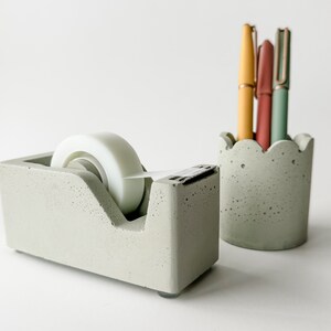 K&ouml;nnte beinhalten: Ein grauer Beton-Klebebandspender mit einer wei&szlig;en Klebebandrolle und ein grauer Beton-Bleistifthalter mit drei Stiften. Die Stifte sind gelb, rosa und gr&uuml;n.