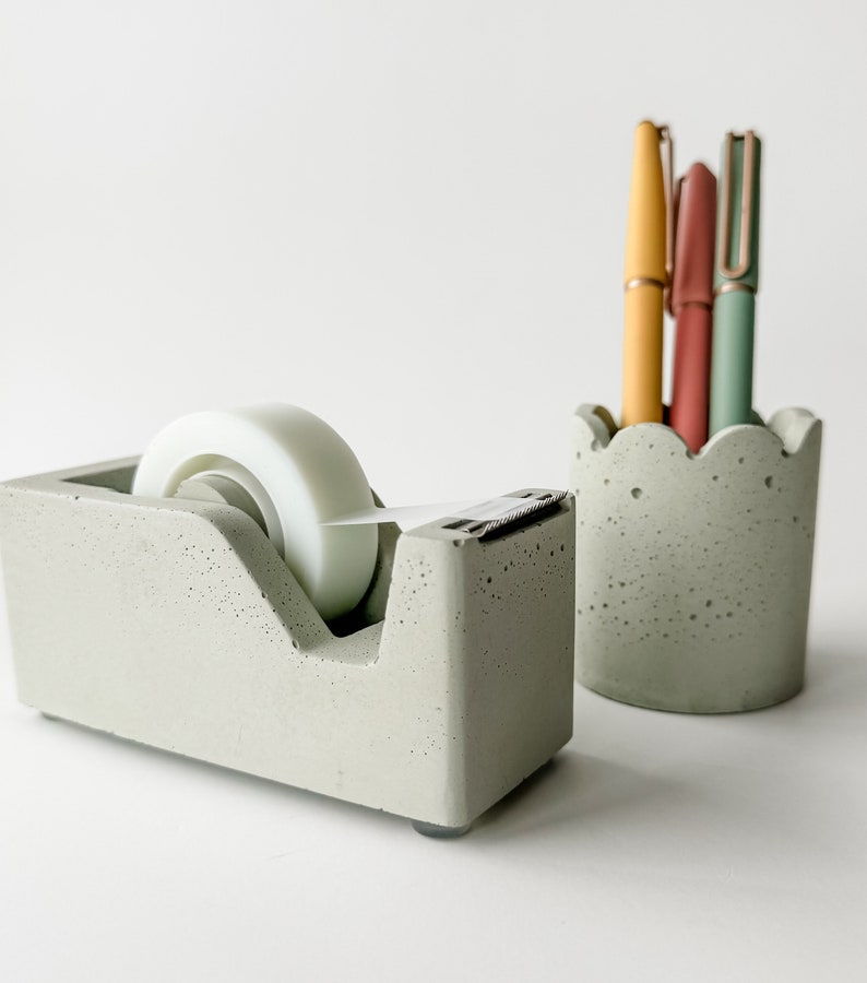 K&ouml;nnte beinhalten: Ein grauer Beton-Klebebandspender mit einer wei&szlig;en Klebebandrolle und ein grauer Beton-Bleistifthalter mit drei Stiften. Die Stifte sind gelb, rosa und gr&uuml;n.