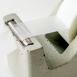 K&ouml;nnte beinhalten: Ein grauer Beton-Klebebandspender mit einer transparenten Klebebandrolle. Der Spender hat eine Metallklinge und eine strukturierte Oberfl&auml;che.