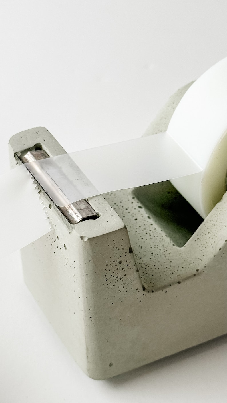 K&ouml;nnte beinhalten: Ein grauer Beton-Klebebandspender mit einer transparenten Klebebandrolle. Der Spender hat eine Metallklinge und eine strukturierte Oberfl&auml;che.