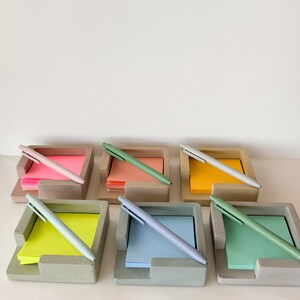 May include: A collection of six concrete desk organizers, each holding a stack of colorful sticky notes and a matching pen. The colors of the sticky notes include pink, orange, yellow, green, and blue. The pens are approximately 14 cm long.