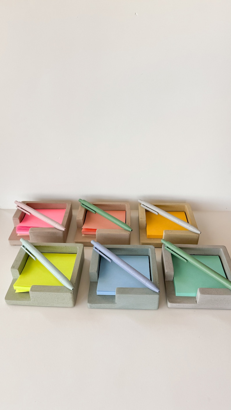 May include: A collection of six concrete desk organizers, each holding a stack of colorful sticky notes and a matching pen. The colors of the sticky notes include pink, orange, yellow, green, and blue. The pens are approximately 14 cm long.