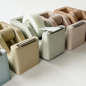 K&ouml;nnte beinhalten: F&uuml;nf Beton-Klebebandspender in verschiedenen Farben, darunter hellblau, beige, gr&uuml;n, hellbraun und braun. Die Spender sind in einer Reihe von links nach rechts angeordnet, wobei der hellblaue Spender links und der braune Spender rechts ist. Ein Klebebandrolle ist im gr&uuml;nen Spender sichtbar.