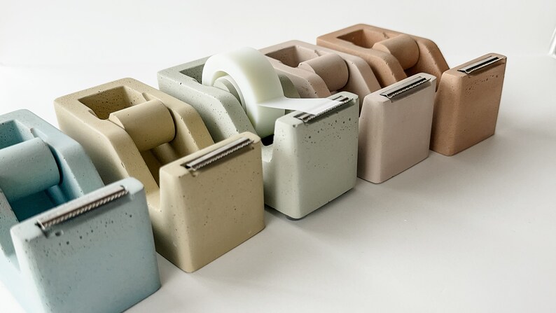 K&ouml;nnte beinhalten: F&uuml;nf Beton-Klebebandspender in verschiedenen Farben, darunter hellblau, beige, gr&uuml;n, hellbraun und braun. Die Spender sind in einer Reihe von links nach rechts angeordnet, wobei der hellblaue Spender links und der braune Spender rechts ist. Ein Klebebandrolle ist im gr&uuml;nen Spender sichtbar.
