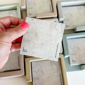 May include: Concrete square coasters in various pastel colors, with a matching square card that says "hi." in a minimalist font. The card is held by a hand with pink nail polish. The coasters have a square frame.