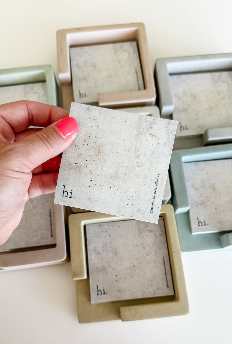 May include: Concrete square coasters in various pastel colors, with a matching square card that says "hi." in a minimalist font. The card is held by a hand with pink nail polish. The coasters have a square frame.