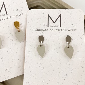 May include: Two pairs of handmade concrete earrings on display. One pair features gold-colored tops and gray teardrop-shaped bottoms. The other pair has dark gray tops and gray teardrop bottoms. The earrings are on white cards with the text "MAVEN HANDMADE CONCRETE JEWELRY".