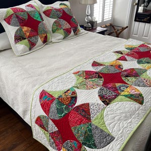 May include: A white bedspread and two matching pillow shams with a colorful floral design. The design features red, green, and patterned fabric in a semi-circular pattern. The bedspread and shams have a quilted texture and a green border.